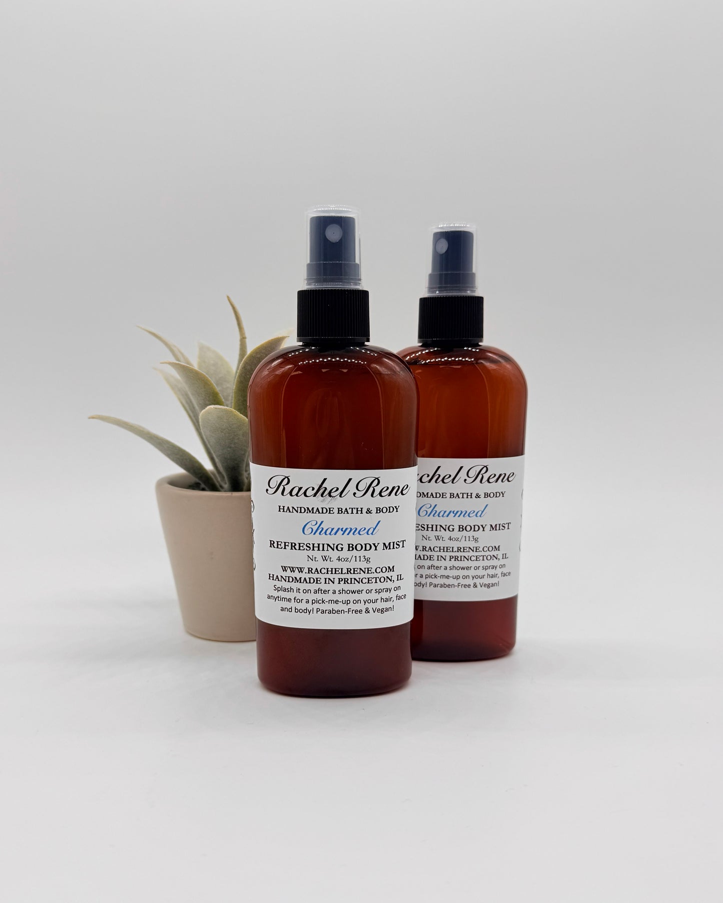 Two brown bottles of Charmed Body Mist with black sprayers and white labels on a gray background.