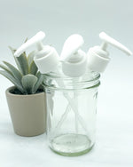 A small glass jar with three white pumps for liquid soap in it. There is a succulent behind it, all on a white background.