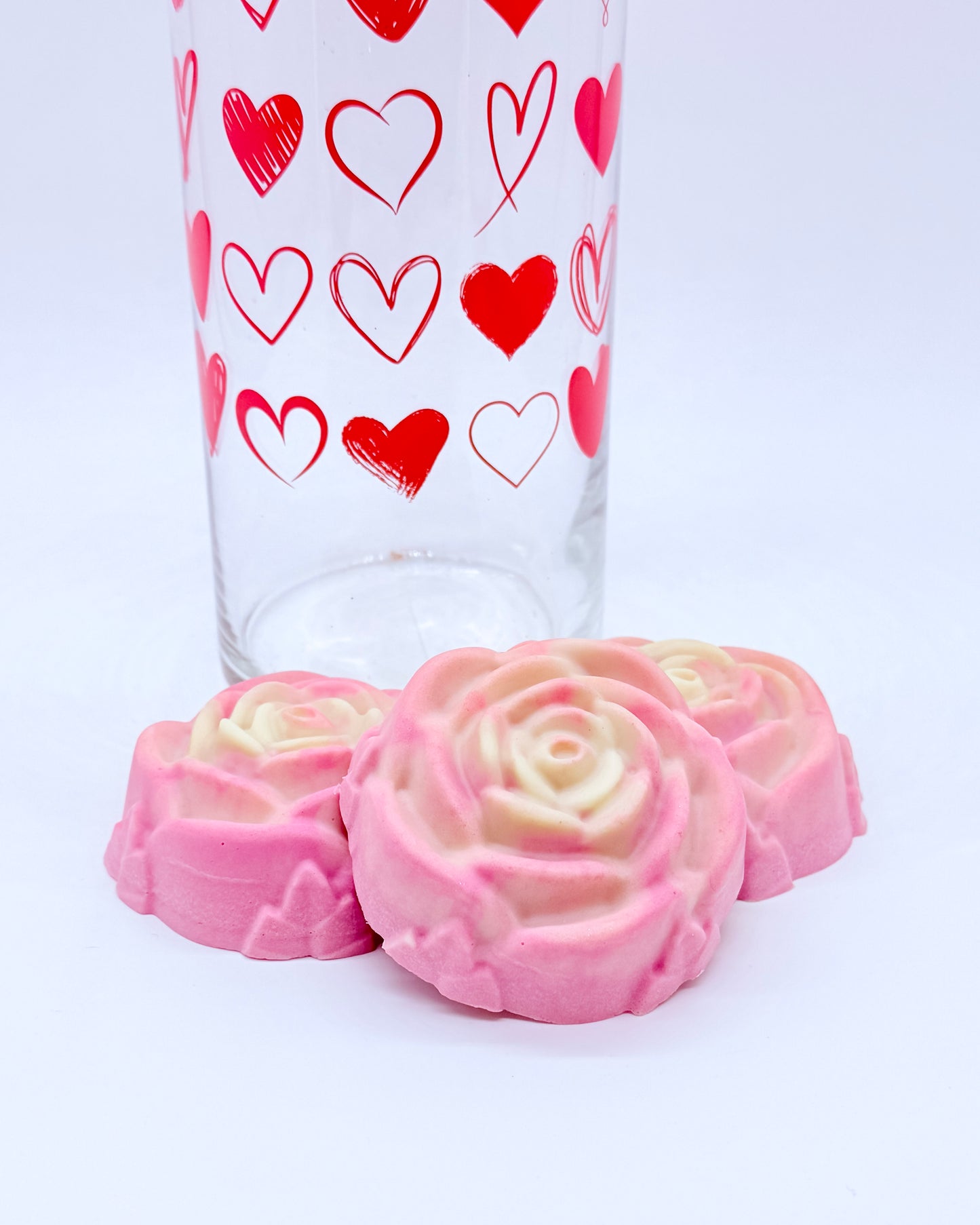 Glass with red heart designs and pink rose-shaped shaving soap bars on a white background