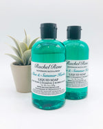 Two semi-transparent teal liquid soaps with white labels reading "Rachel Rene: Handmade Bath & Body - Aloe & Summer Herbs. Liquid Soap - Body Wash - Shampoo - Bubble Bath." They have black pull-top caps and stand in front of a potted succulent and white background.