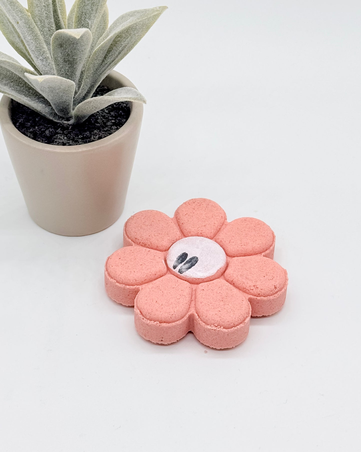 Pink flower-shaped shower bomb with white center on a white surface, next to a small potted plant.